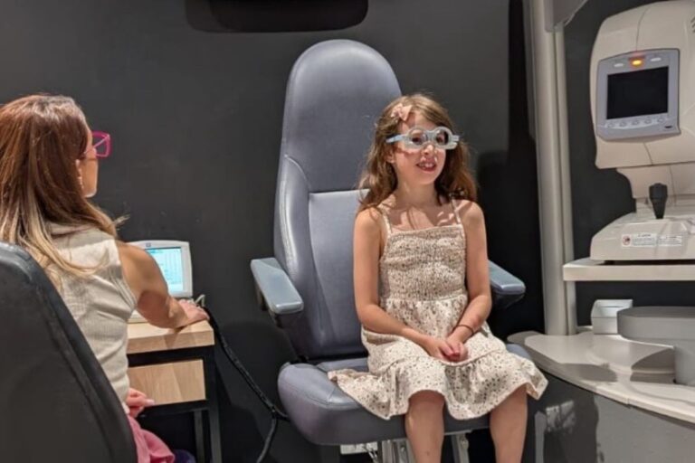 A young girl sitting in an eye exam chair, looking curiously at the eye doctor in a bright examination room.