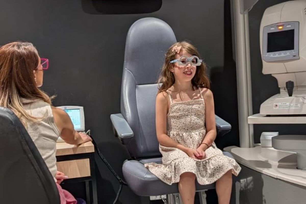A young girl sitting in an eye exam chair, looking curiously at the eye doctor in a bright examination room.