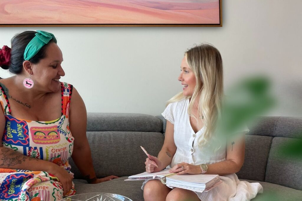 Two women sitting on a couch, engaged in a conversation with friendly expressions.