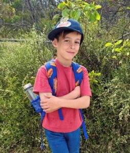 A boy in a cap and red shirt carries a backpack on his shoulders. 