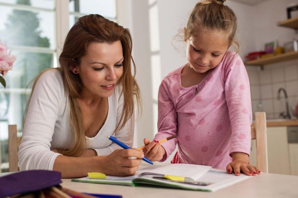 A woman assists a young girl with a project, both concentrating on their work at a table filled with supplies.