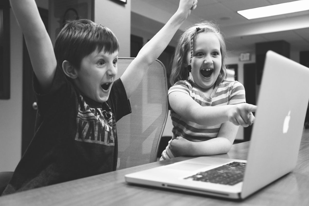 Two children sit together in front of a laptop, engaged in an activity on the screen.