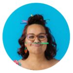 A woman with a moustache playfully holds a pencil in her mouth, looking thoughtful and engaged in her work.