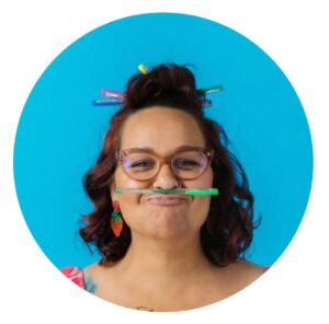 A woman with a moustache playfully holds a pencil in her mouth, looking thoughtful and engaged in her work.