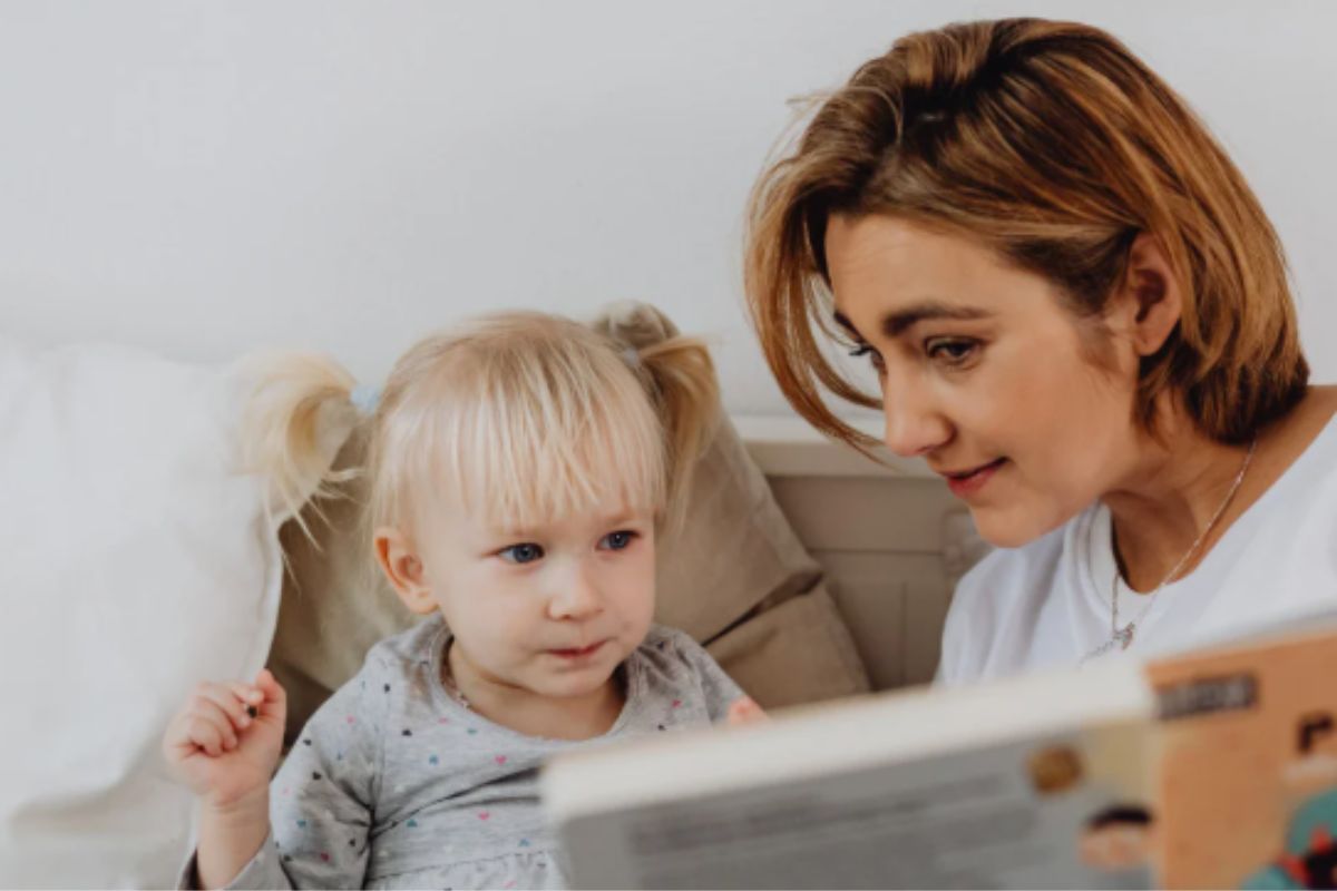 A woman reads a book to a toddler in bed.