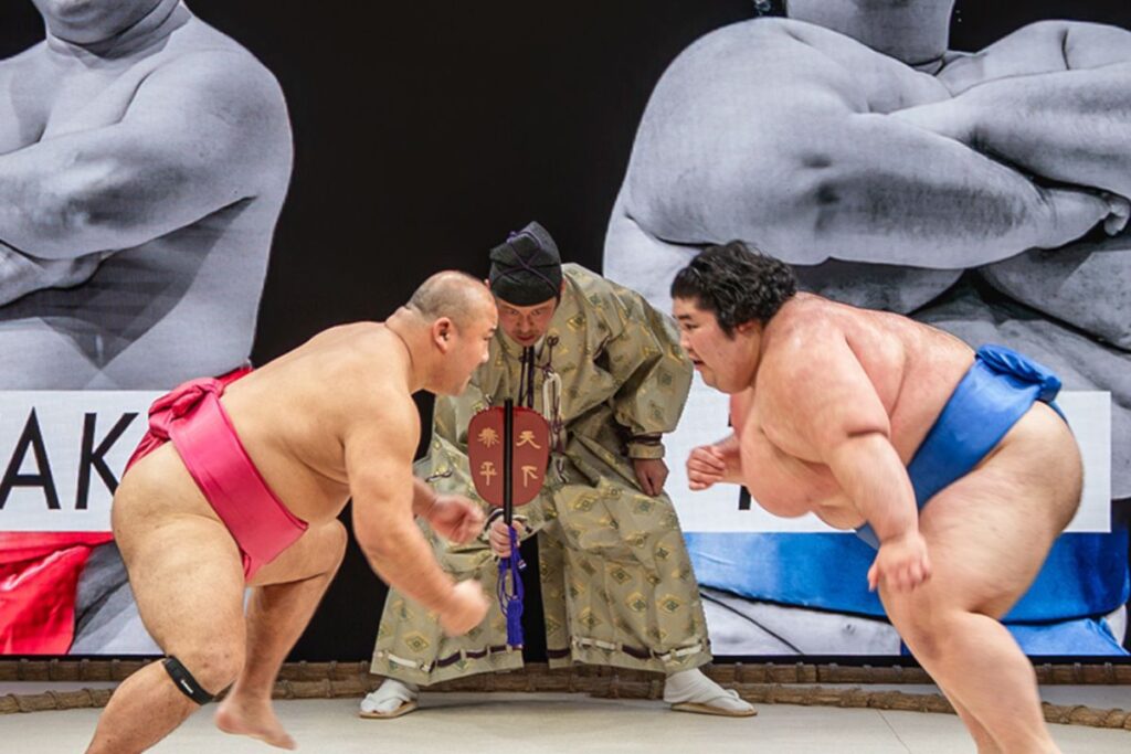 Two sumo wrestlers face off in the ring, while a large screen shows event details behind them.