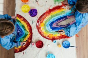 Two children paint rainbows on a colourful artful table with pot paints.