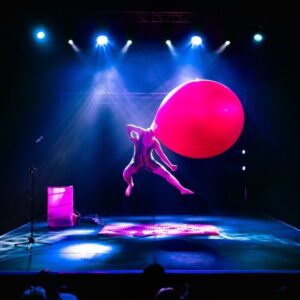 A man performs acrobatics on a stage with a giant balloon for a head.