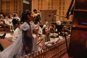 A young girl in a dress looks on in awe at the cello as an orchestra play to a crowd of children.