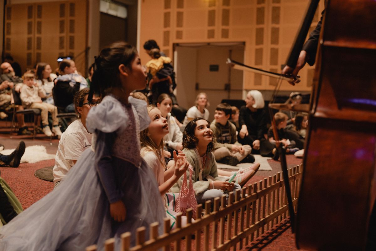 A young girl in a dress looks on in awe at the cello as an orchestra play to a crowd of children.