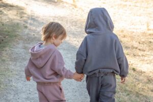 two children walking on a dirt road holding hands dressed in hooded jumper and pant sets.