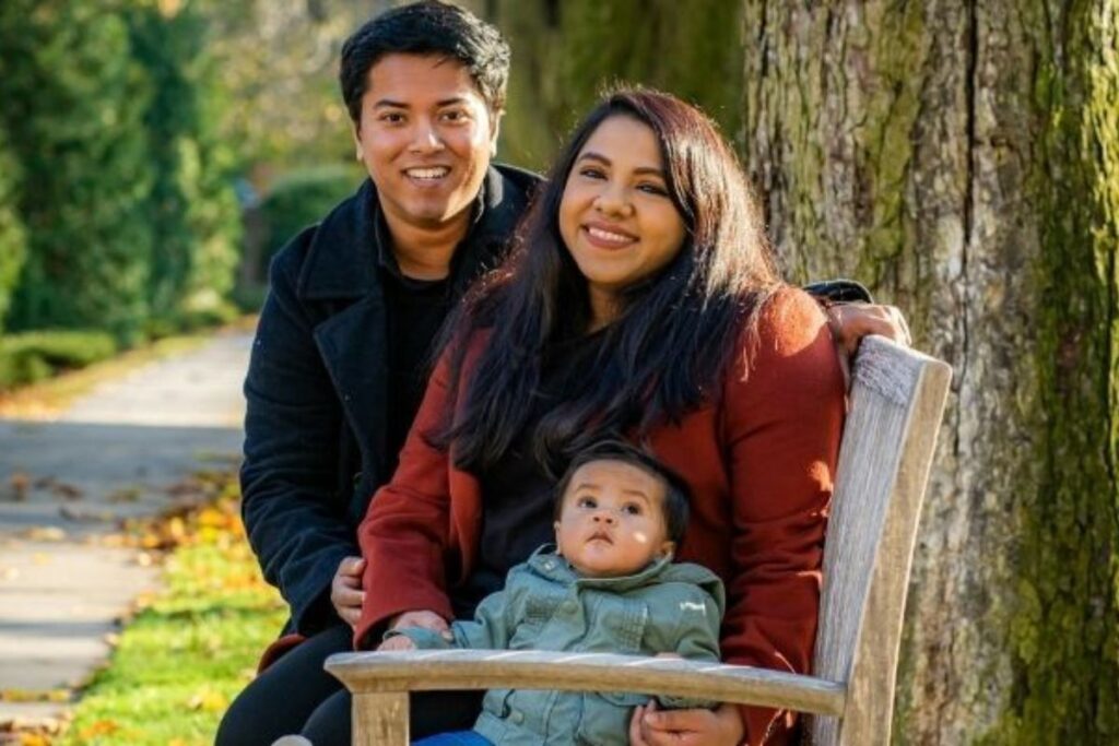 A young family sit together on a park bench dressed in warm clothing surrounded by trees and autumn leaves.