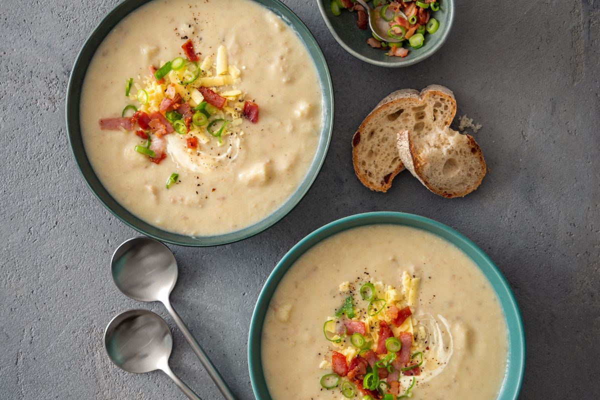 2 bowls of potato and leak soup with colourful garnish and bread and 2 spoons on a table.