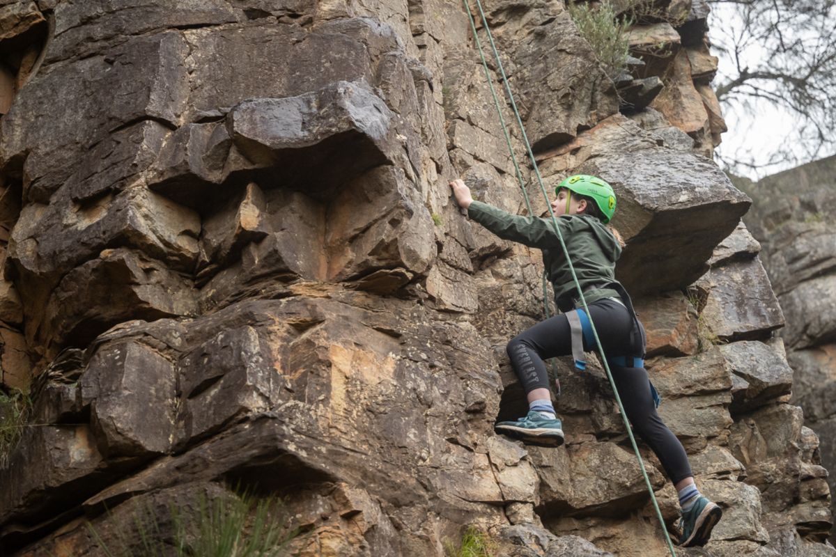 A child wearing a helmet and harness scales a natural rock face on a roped climbing adventure