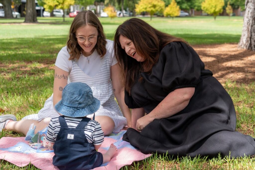 Two women and a baby sat on a blanket on green grass in a park.