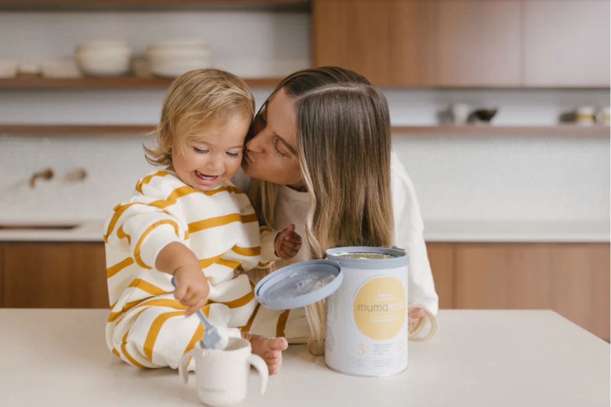 Mother sitting on a kitchen floor with her baby beside mumamoo infant formula tins on a wooden surface