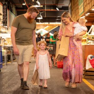 A family with two young children walking through Adelaide Central Market, carrying shopping bags past market stalls.