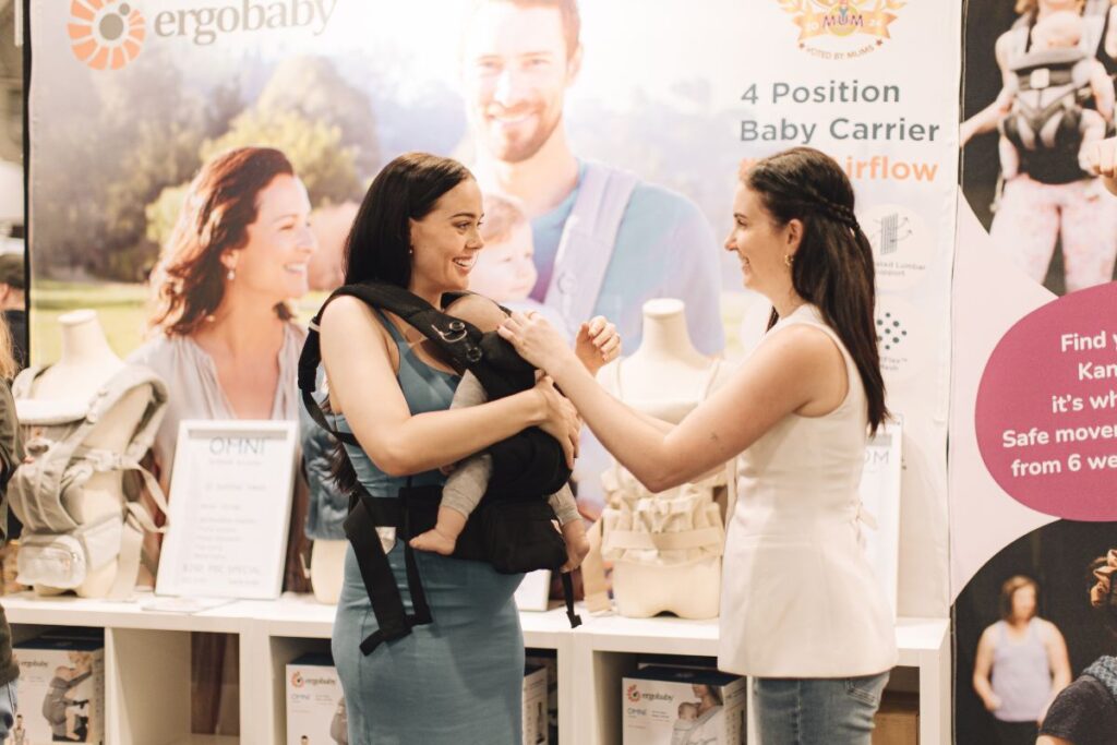 A woman wearing a black Ergobaby baby carrier with a newborn inside chats and smiles with another woman at a baby expo, with Ergobaby product displays and signage visible in the background.