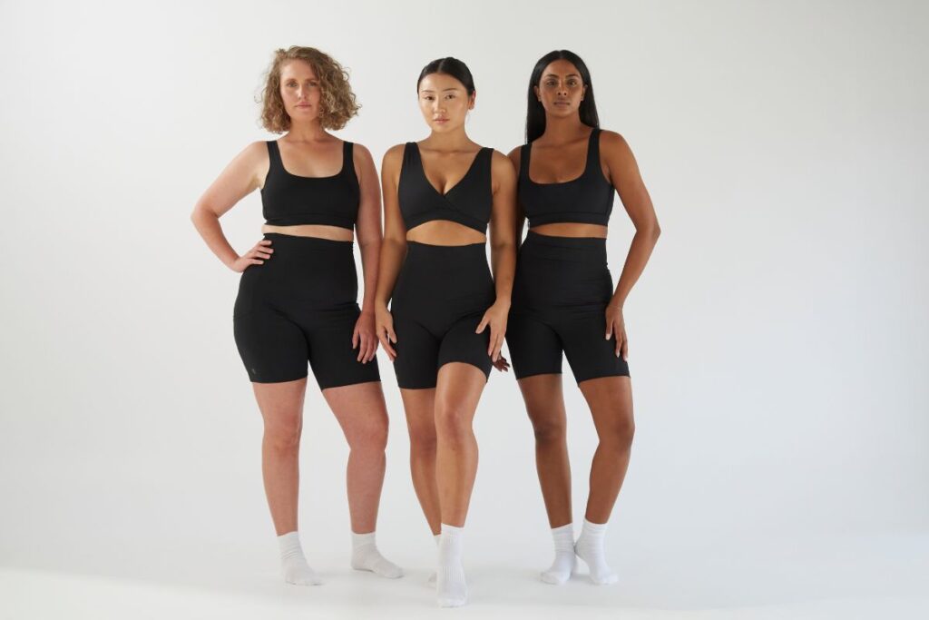 Three women of different body types wearing Bare Mum black postpartum recovery bralettes and bike shorts against a white background