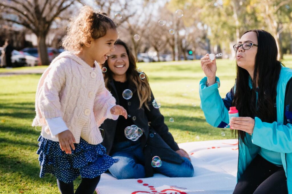 Two ladies and a toddler sitting on a rug outdoors playing with bubbles and smiling.