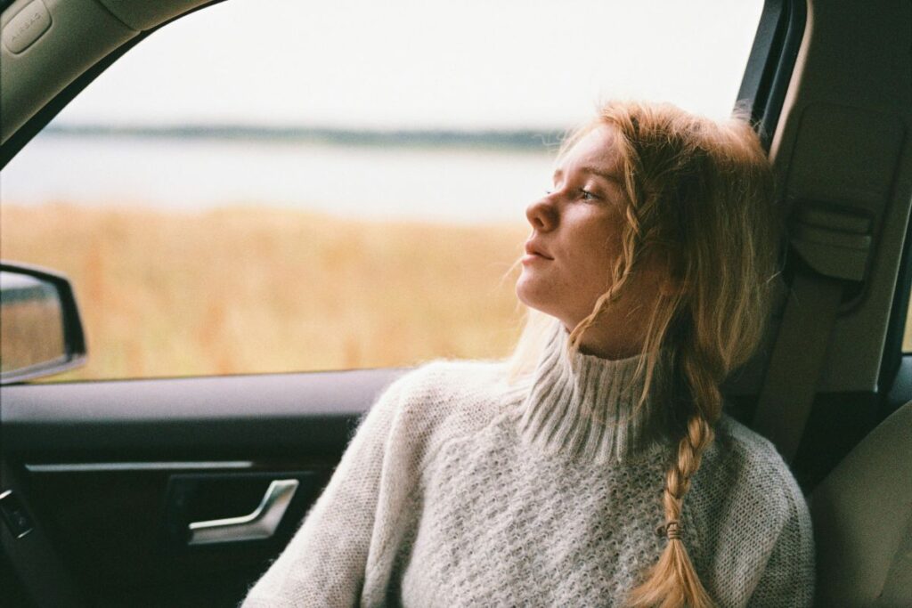 A young woman with a long blonde braid sits in the passenger seat of a car, gazing thoughtfully out the window toward an open landscape.