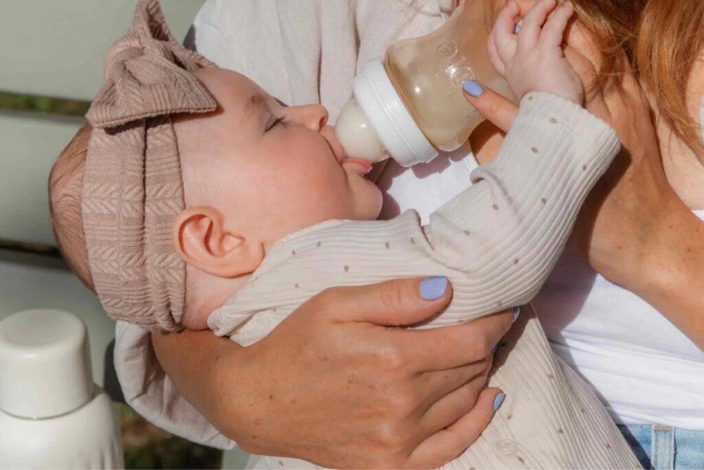 An infant wearing a bow headband being bottle fed by mother with green painted nails.
