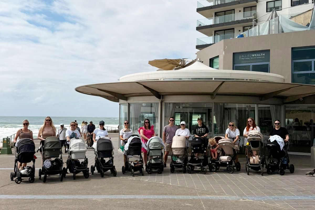 A group of mums with prams gathered in a beachside carpark preparing for a walk together.