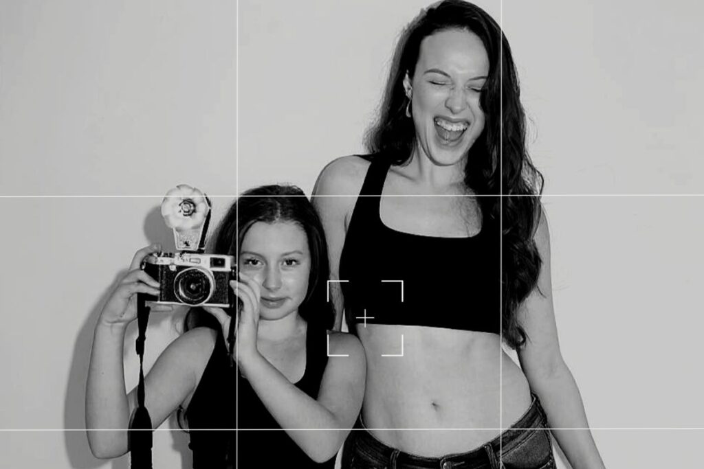 A black and white campaign image featuring a young girl holding a vintage camera alongside an older model wearing a Billie Green black crop top, laughing together in a studio setting.