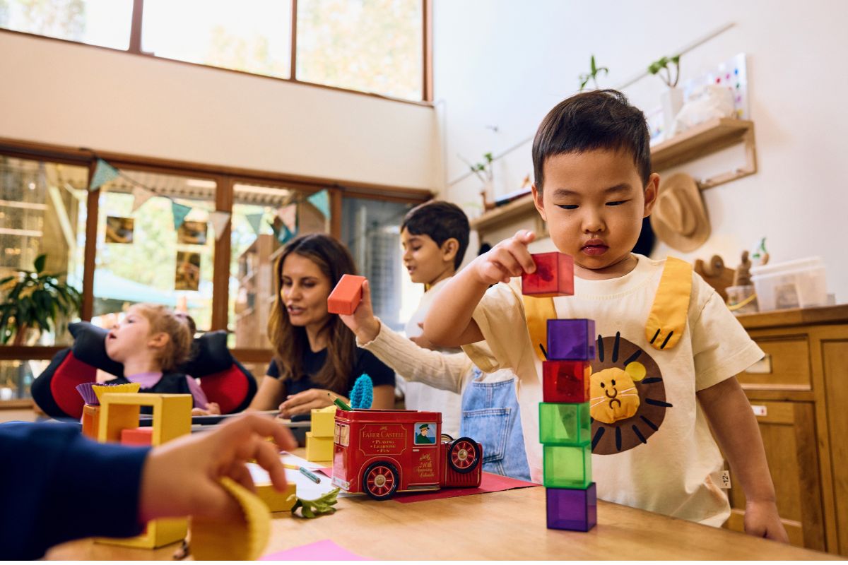 A young child stacks colourful blocks at a preschool activity table with a teacher and other children nearby