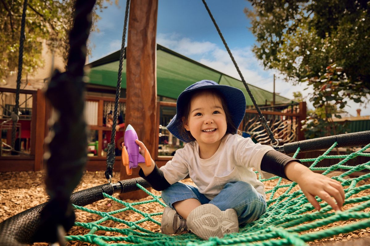 A smiling young child sits on an outdoor rope swing holding a toy rocket at a preschool playground
