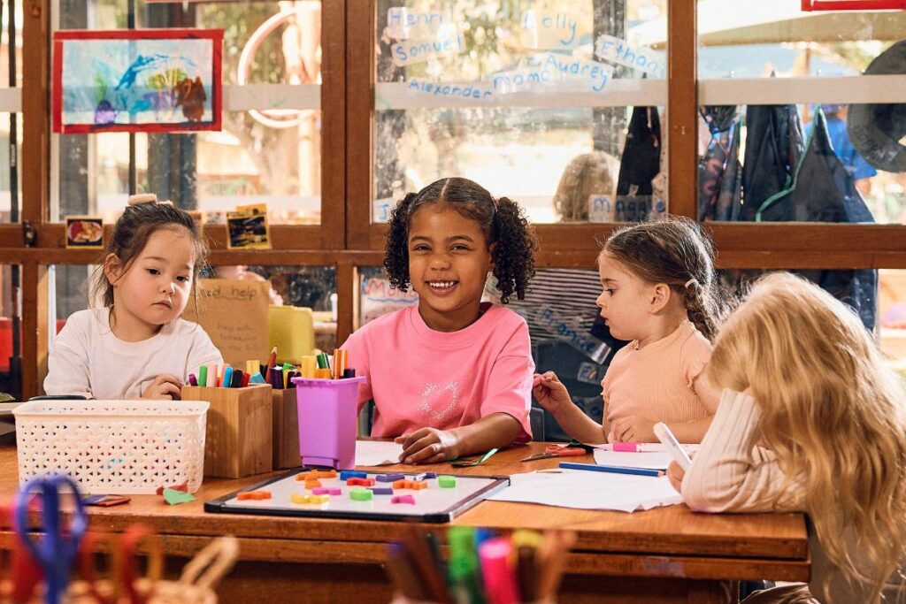 A group of preschool children sit together at a classroom table engaged in creative play and learning activities