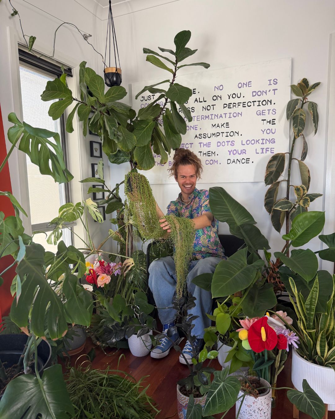 Mr Luke smiling among his houseplant collection, holding a trailing string of pearls plant in his lounge room
