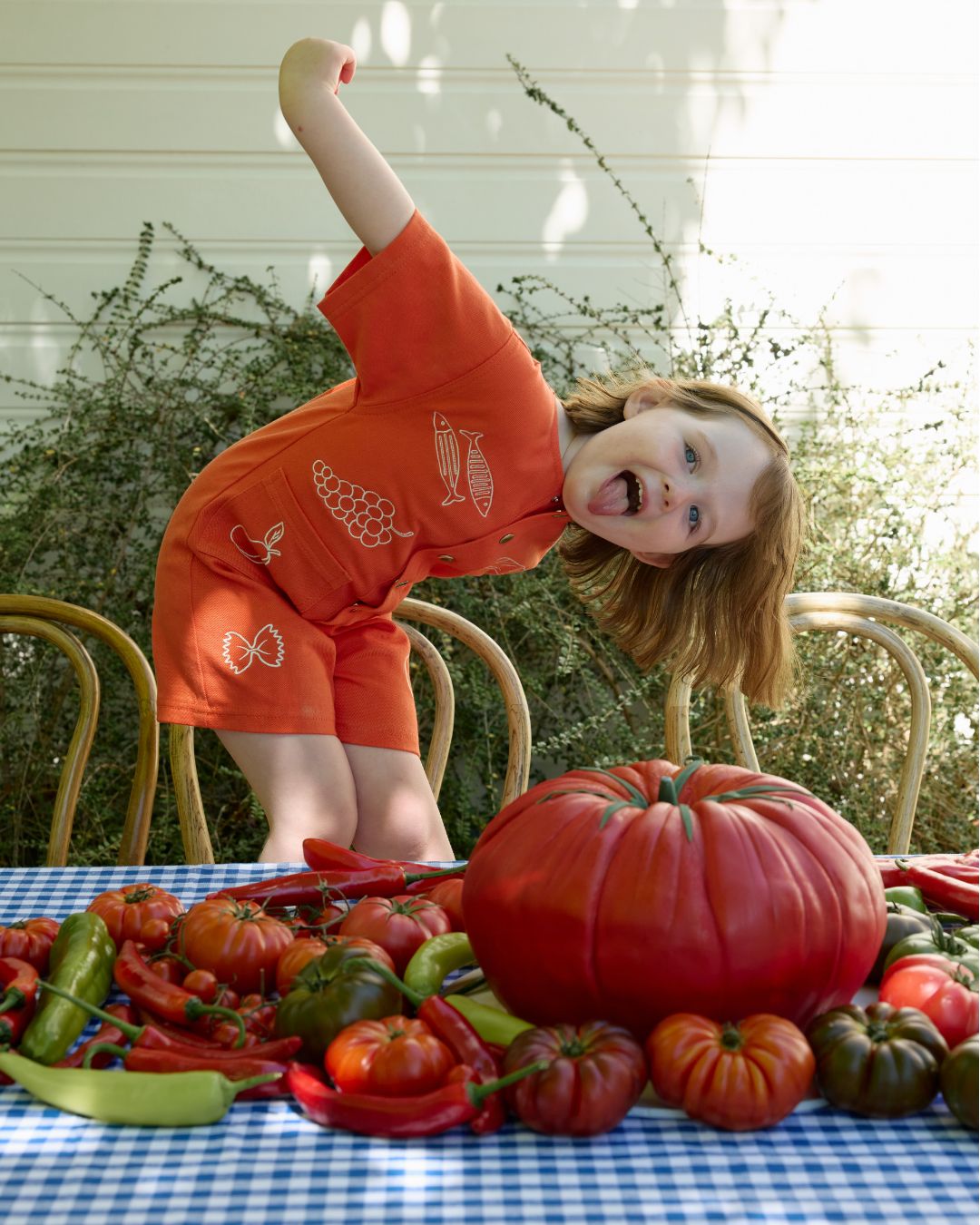 A girl wearing a red playsuit with printed sardines, grapes and pasta playfully pokes her tongue out with a table arrangement of garden vegetables in front of her.