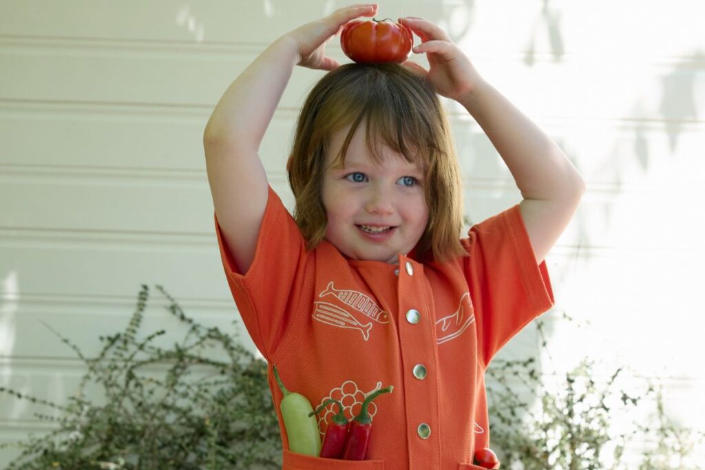 A girl wearing a red playsuit with printed sardines, grapes and pasta joyfully holds a tomato over head with a table arrangement of garden vegetables in front of her.