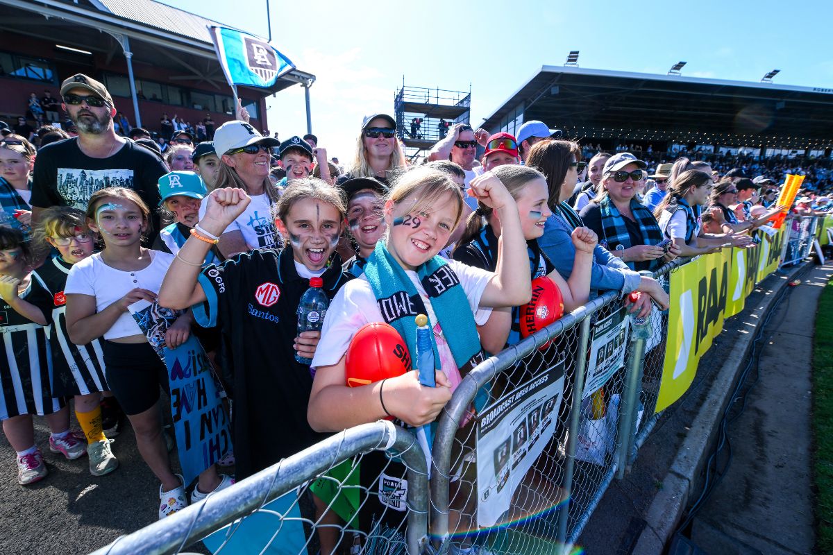 Excited young Port Adelaide fans with face paint and scarves cheering at an AFLW home game at Alberton Oval