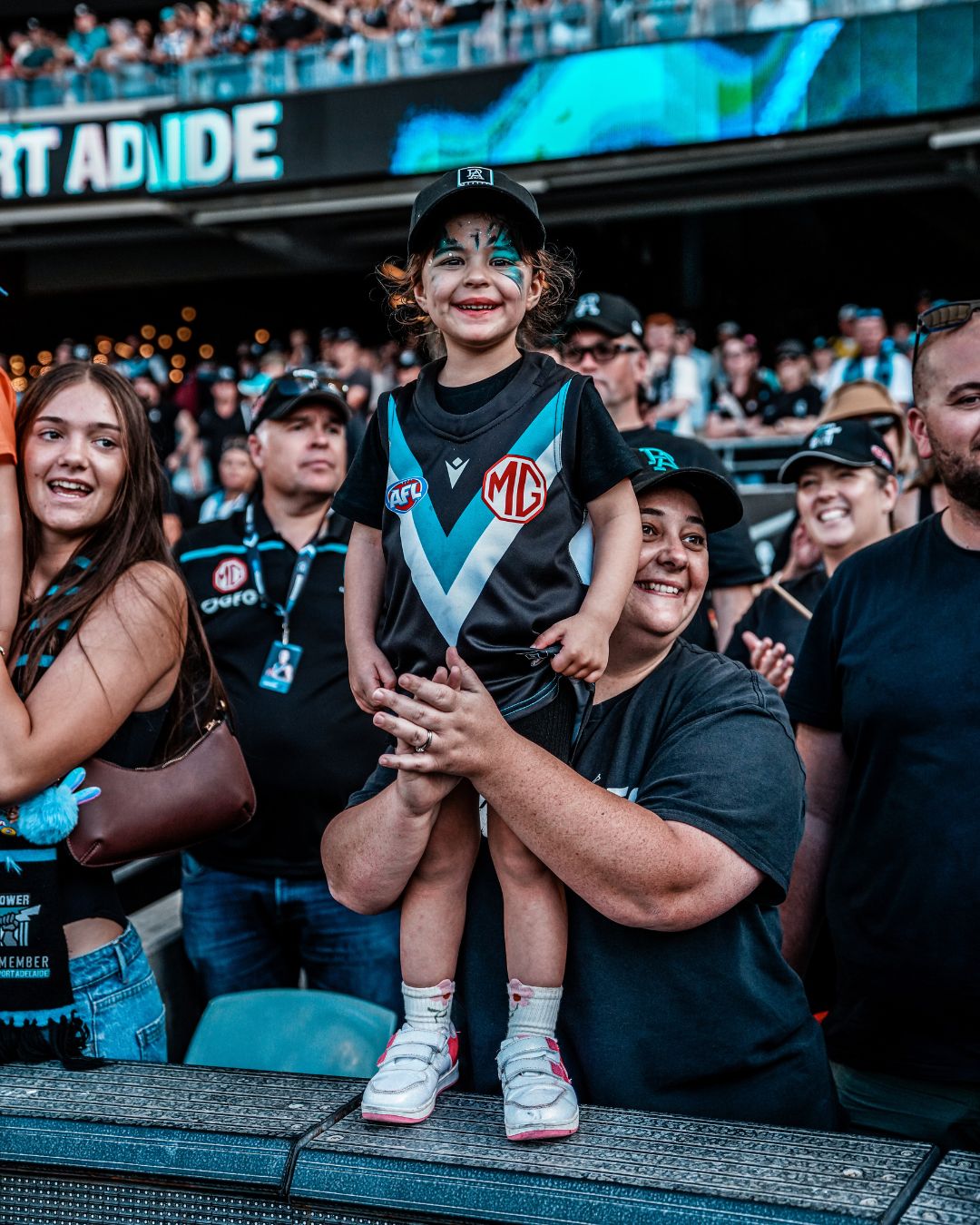 Young Port Adelaide fan in team guernsey and face paint held up by a family member in the crowd at Adelaide Oval
