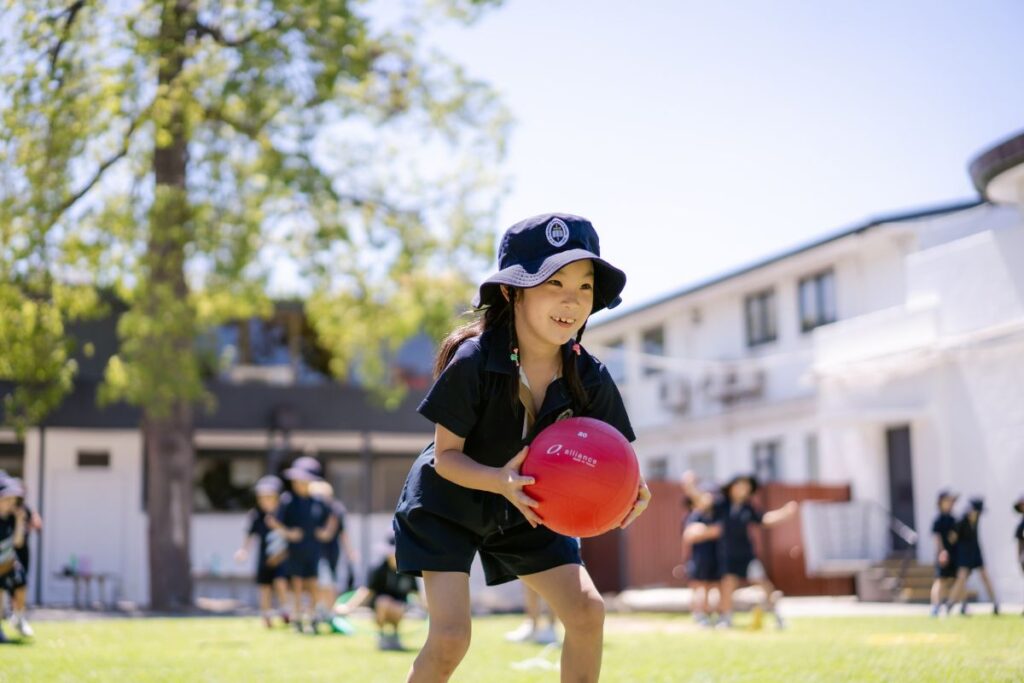 A young Pulteney Grammar Reception student in school uniform and sunhat playing with a red ball on the school grounds