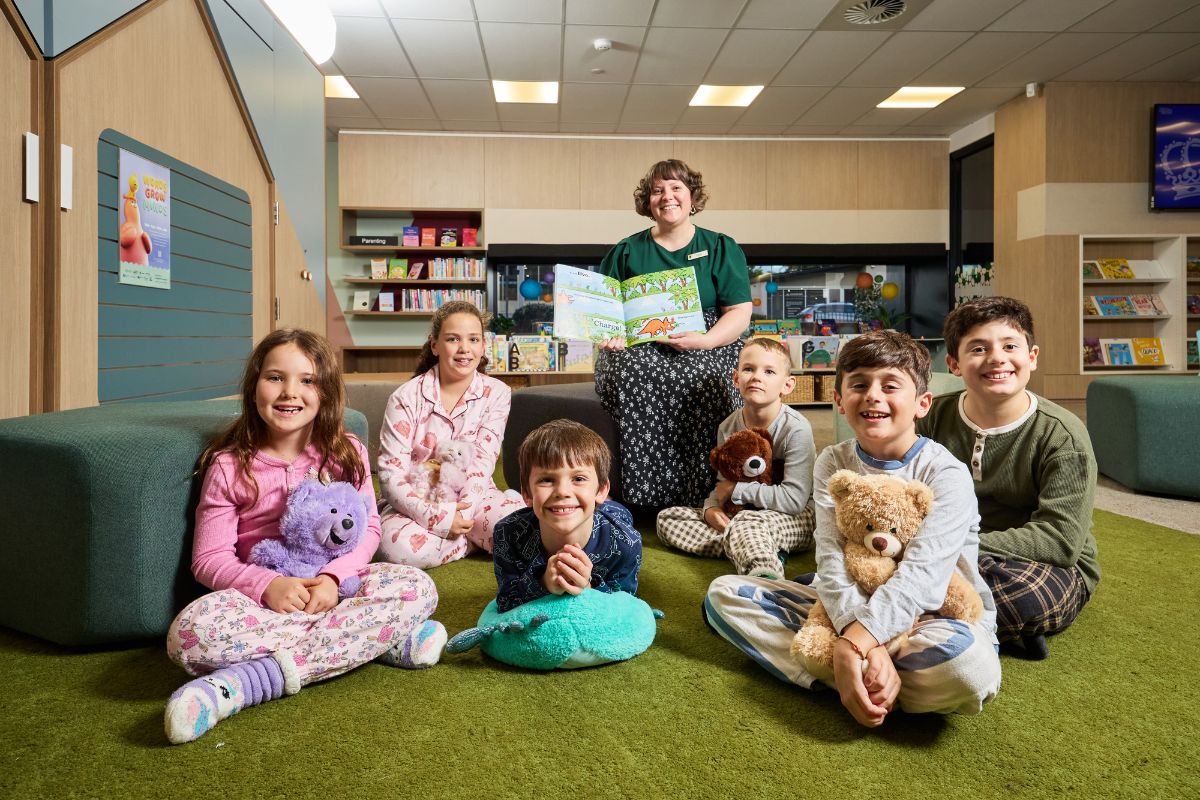 A librarian in a green shirt reads aloud to six primary school-aged children in pyjamas, who sit on a green carpet holding stuffed animals in a library setting.