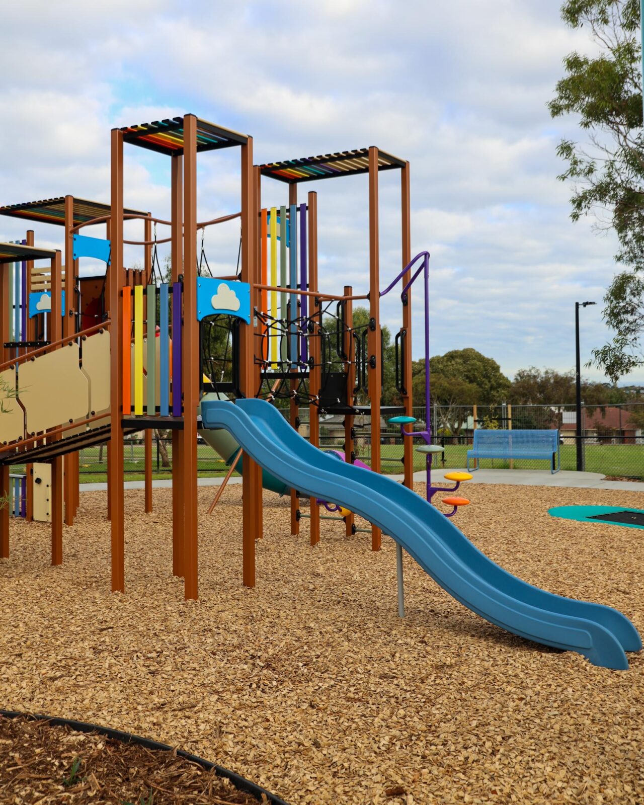 The multi-level play structure at Southbank Boulevard Reserve playground in Sheidow Park, featuring a double blue slide, climbing elements and a wheelchair-accessible trampoline on a wood chip surface.