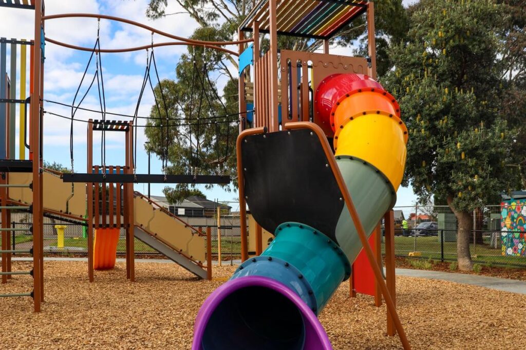 A colourful rainbow tunnel slide at the newly upgraded Southbank Boulevard Reserve playground in Sheidow Park, with a climbing structure and trees visible in the background.