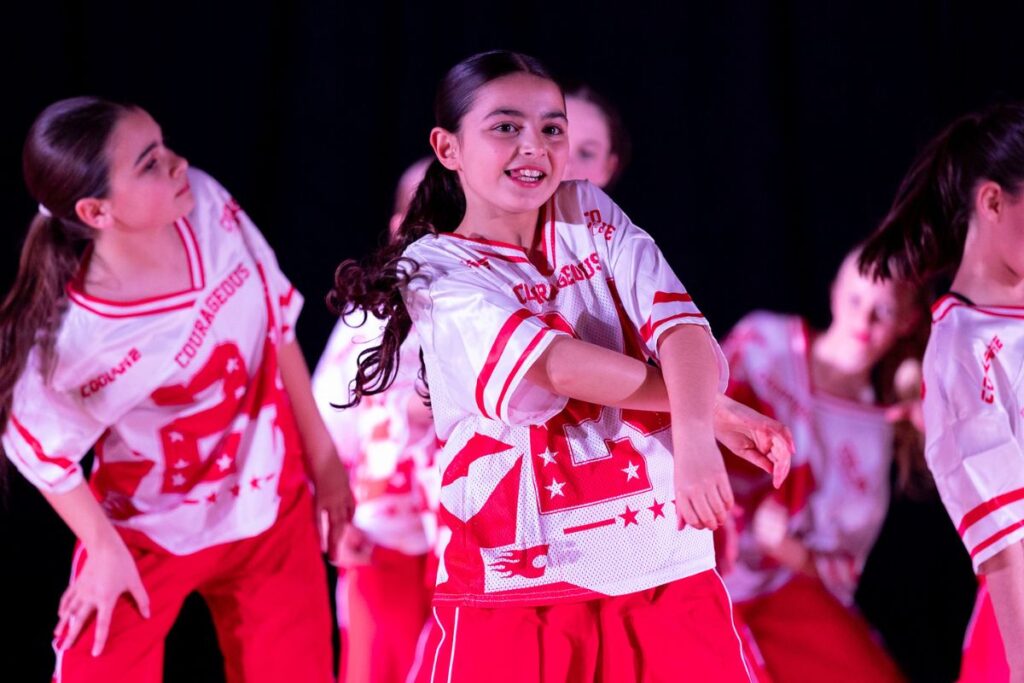 Students perform on stage in red and white costumes during a school dance showcase.