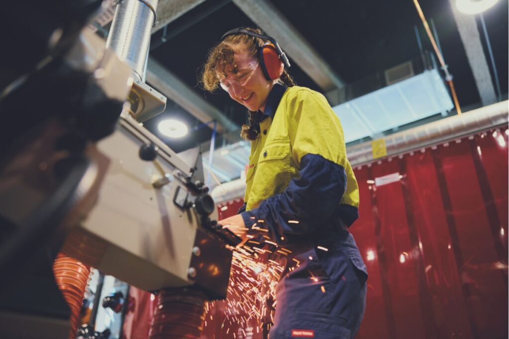 A female SA Technical College student in hi-vis workwear and safety gear operating machinery with sparks flying in a workshop.