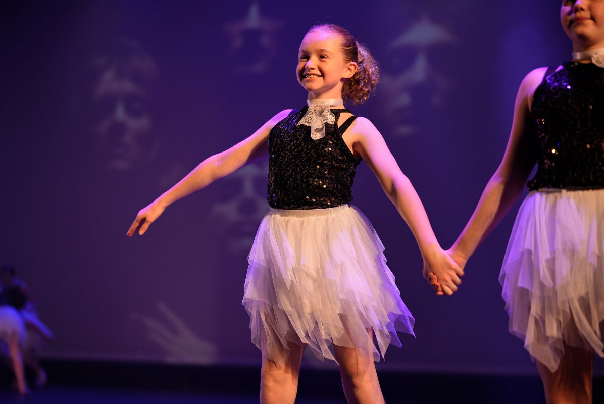 A young girl smiles while performing on stage in a dance recital costume of black sequins and white tulle skirt.