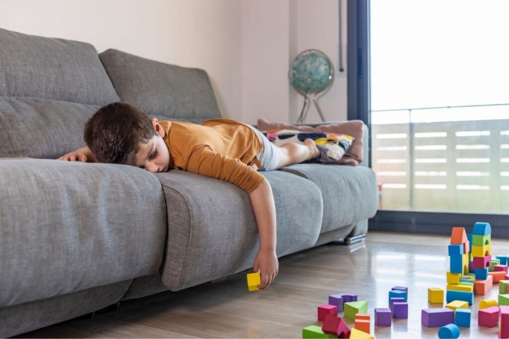 A young boy lies face down on a couch, arm dangling to reach colourful building blocks scattered on the floor below.