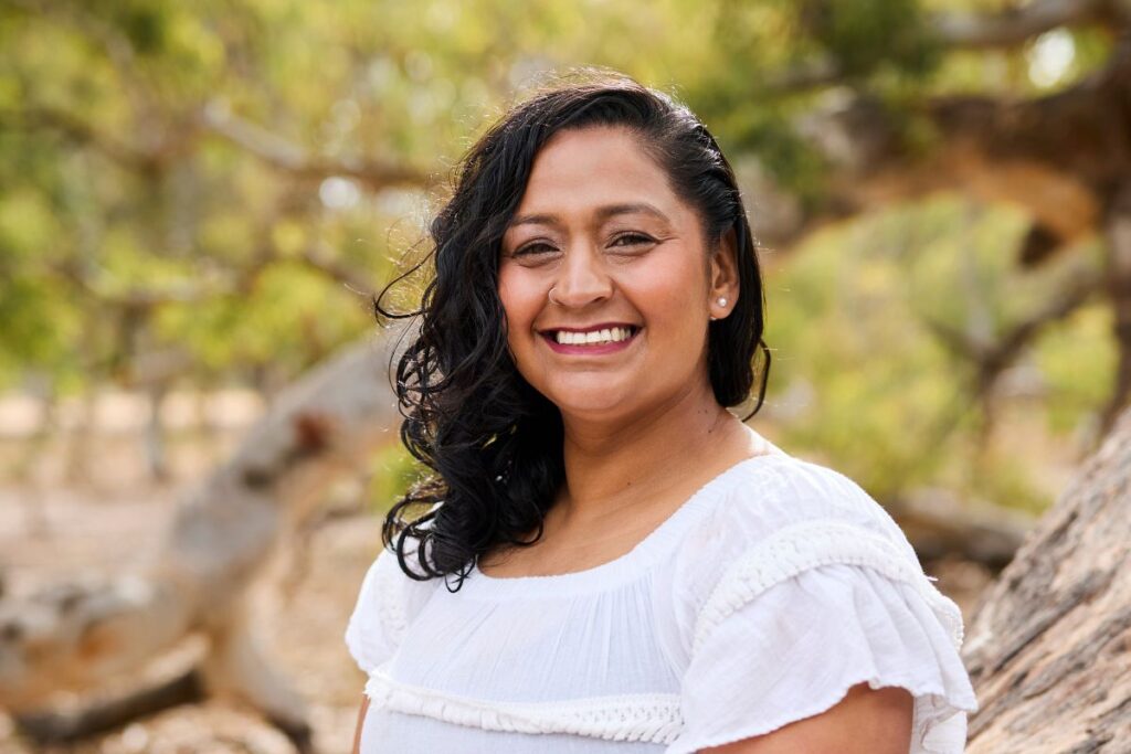 Rochell Toia, founder of Cedarwood Road, smiling outdoors in a white top with green foliage in the background.