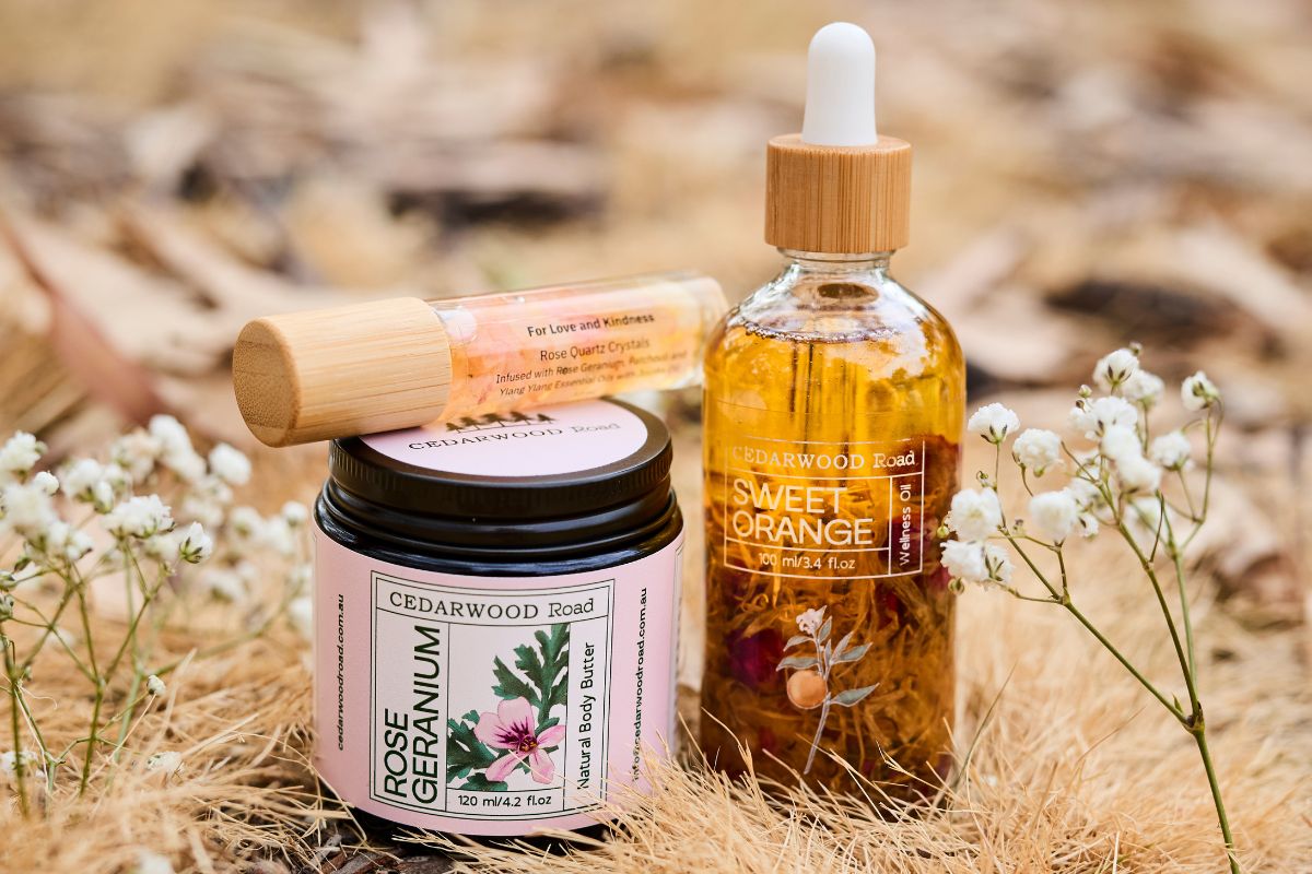 Cedarwood Road Rose Geranium natural body butter and Sweet Orange wellness oil, with a rose quartz crystal roll-on, styled on dried grass with baby's breath flowers.