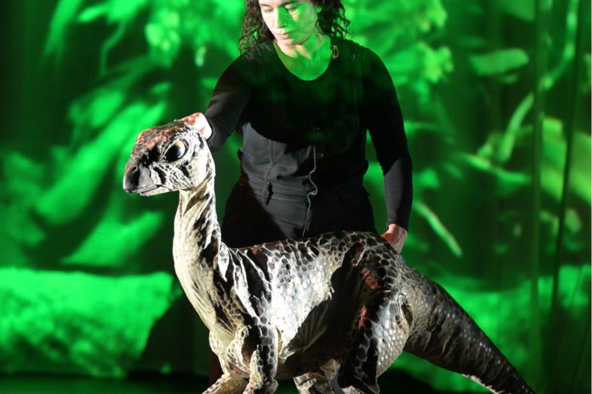 A scaly dinosaur puppet lit in green stage light in Erth's Dinosaurs