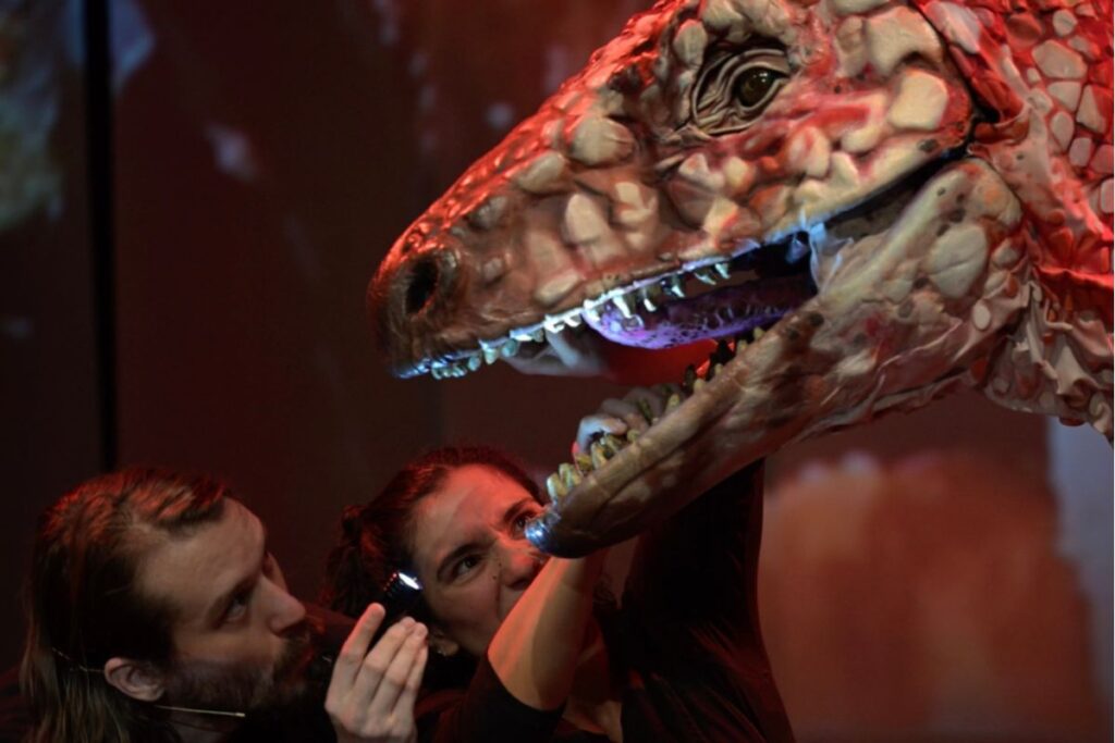 Close-up of a life-sized dinosaur puppet's head being operated by puppeteers in Erth's Dinosaurs