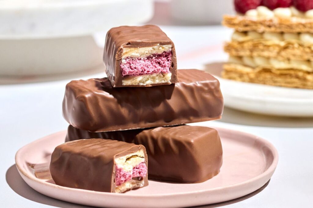 The Haigh's Milk Raspberry Mille Feuille Bar cut in half to reveal its layered raspberry and white chocolate ganache centre, stacked on whole bars with mille feuille pastry and fresh raspberries in the background
