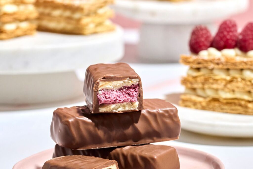 The Haigh's Milk Raspberry Mille Feuille Bar cut in half to reveal its layered raspberry and white chocolate ganache centre, stacked on whole bars with mille feuille pastry and fresh raspberries in the background